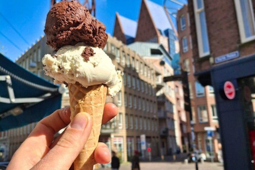 A hand holding an ice cream cone with two scoops, one chocolate and one vanilla, in an urban setting with buildings in the background. 2