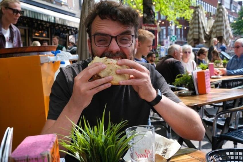 A person eating a sandwich at an outdoor cafe with tables and chairs, surrounded by other people and greenery. 1
