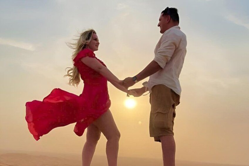 A couple holding hands, a woman in a red dress, and a man in a white shirt, standing on a beach with the sun setting in the background. 2