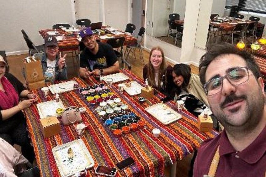 A group of people seated around a table with various items and a colorful tablecloth. 5