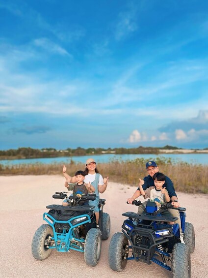 Vier Personen fahren mit Quads auf einem sandigen Pfad, im Hintergrund ein Gewässer und Vegetation. 1
