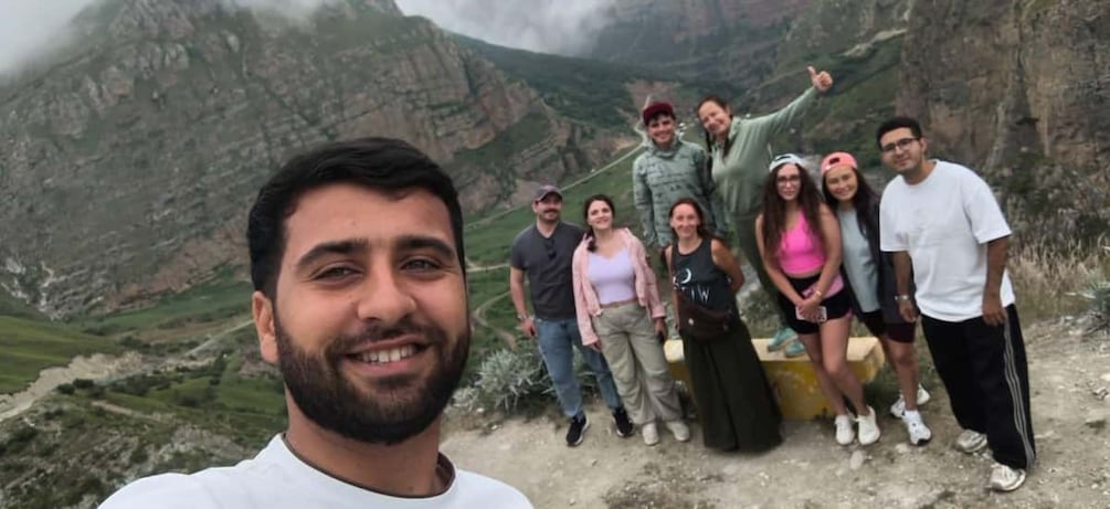 A group of people standing on a mountain trail with rocky terrain and greenery in the background. 5