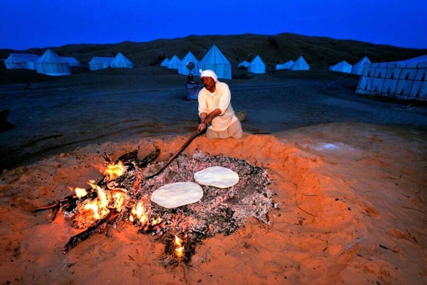 Una persona cocina sobre fuego abierto en un paisaje desértico, con tiendas de campaña al fondo. 3