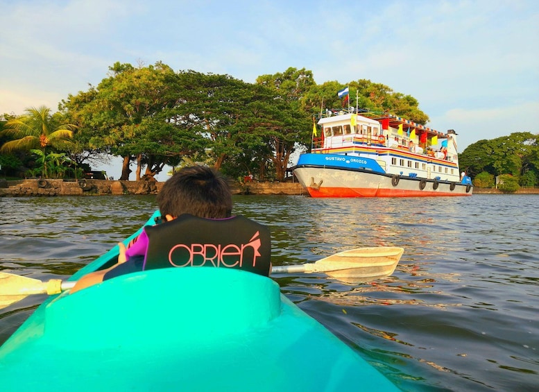 Una persona en un kayak, con chaleco salvavidas marcado 'OBRIEN', rema sobre el agua. Un barco grande y colorido llamado 'GUSTAVO OROZCO' está amarrado al fondo. 3
