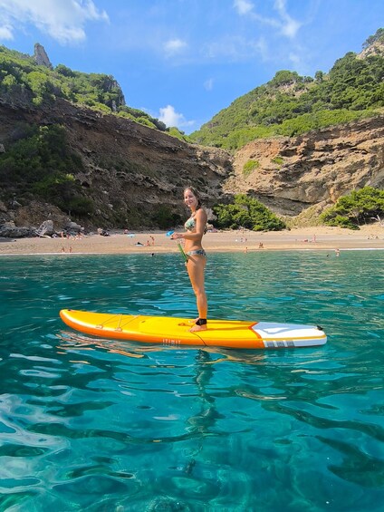 Una persona de pie en una tabla de remo amarilla en aguas azul cristalina, con una costa rocosa y vegetación al fondo. 2