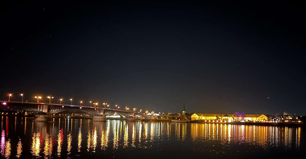 A nighttime view of a lit bridge over a body of water with reflections of lights on the water's surface. 1