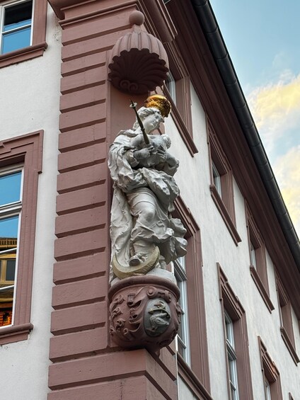 A statue of a robed figure holding a child, mounted on a column with ornate carvings, set against a building facade with windows and a clear sky. 4