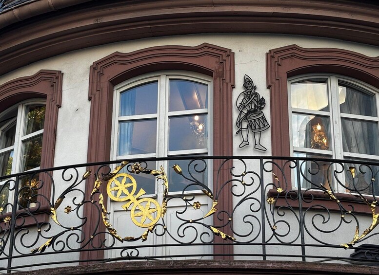 A building facade with ornate window frames, decorative metal railings, and a relief sculpture of a figure playing a drum. 2