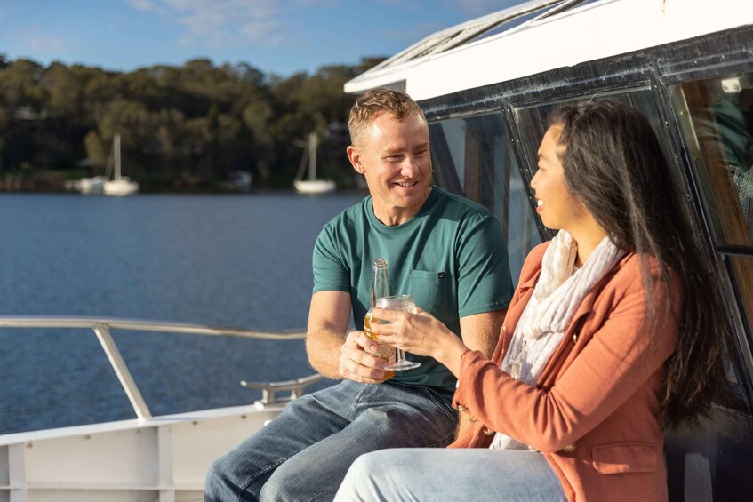 Two people sitting on a boat, holding drinks, with a lake and boats in the background. 2