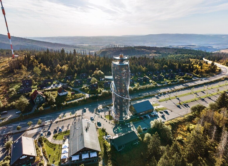 A modern tower with a spiral staircase, surrounded by a parking lot and residential buildings, with a forested landscape in the background. 3