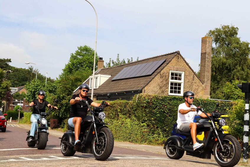 Tres personas montando scooters eléctricos por una calle con una casa de ladrillo y follaje en el fondo. 1