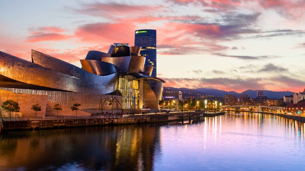 A modern architectural building with a unique design, a river in the foreground, and a cityscape in the background under a pink sky. 2