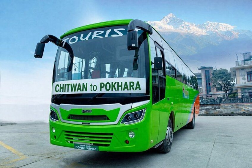 A green bus with 'CHITWAN to POKHARA' written on the front, parked on a paved surface with mountains in the background. 2