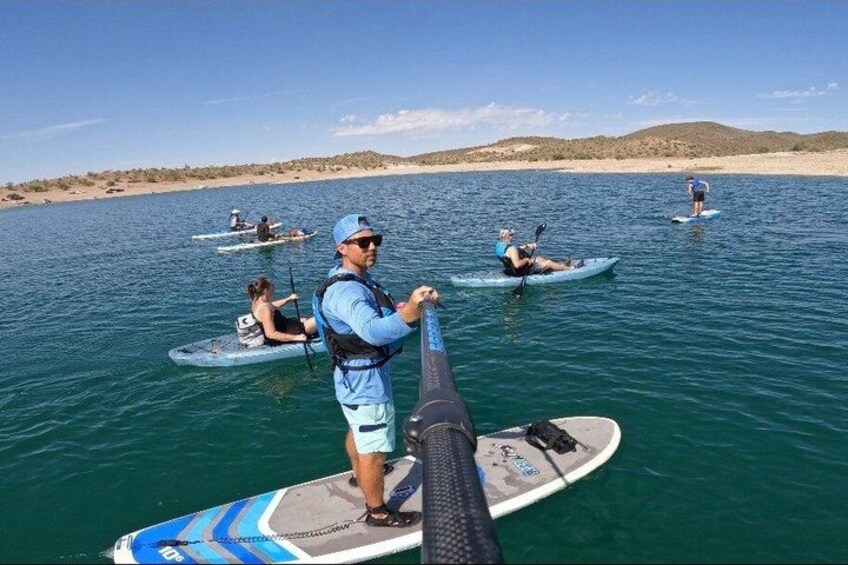 People paddleboarding on a calm body of water with a sandy beach and hills in the background. 2