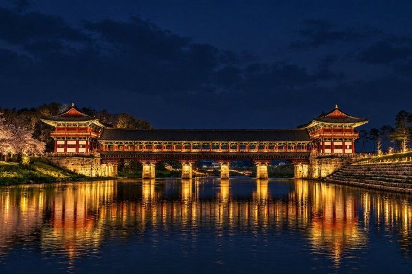 A traditional Korean bridge with a reflection in the water, surrounded by trees and illuminated buildings. 2