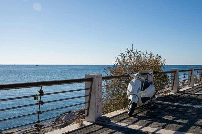 A white scooter parked on a paved walkway by the sea, with a railing and a clear view of the water. 4