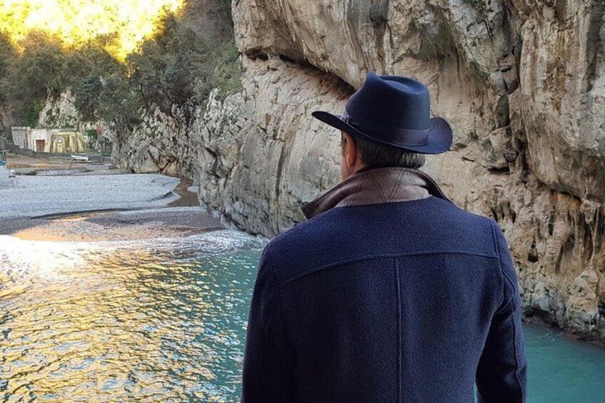 A person in a hat and coat standing by a rocky riverbank with a building in the background. 2