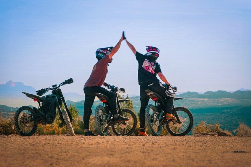 Two people on motorcycles, one giving a high-five to the other, with a scenic mountain landscape in the background. 1
