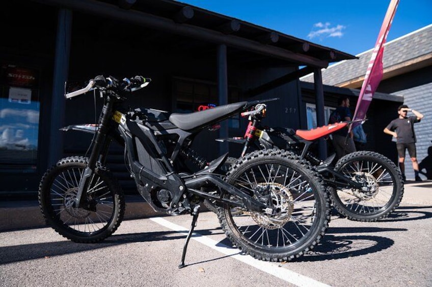 Two dirt bikes parked in a lot with a building in the background. 2