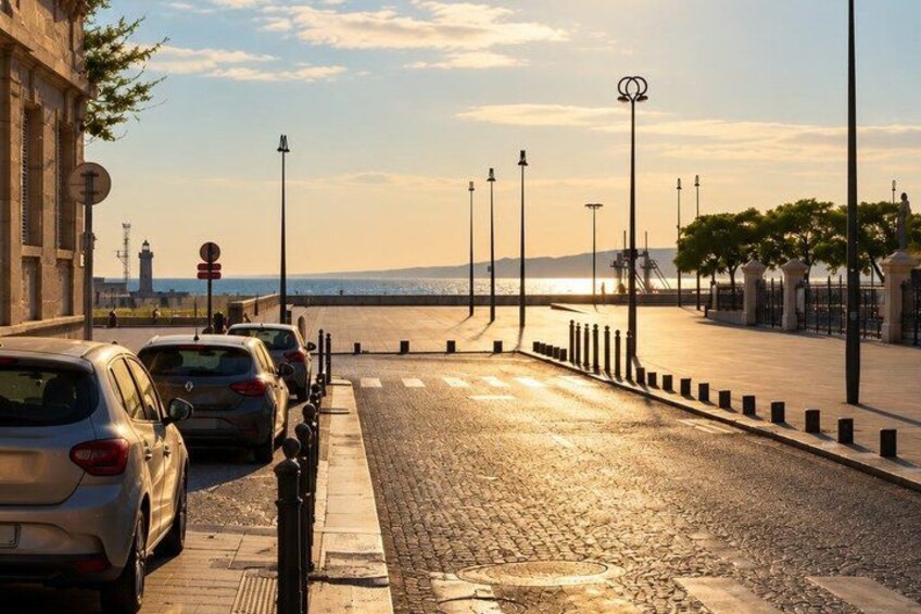 A coastal road with parked cars, a paved walkway, street lamps, and a distant view of the sea. 3