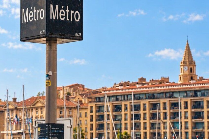 A metro sign with the word 'Metro' in two languages, a clock tower, and a building with a red roof. 5