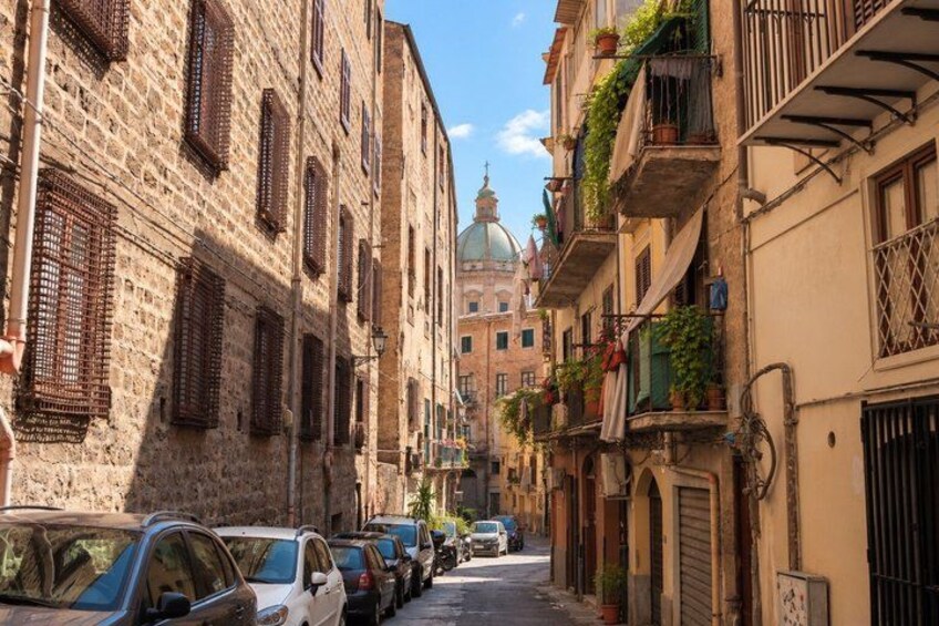 A narrow street lined with historic buildings, parked cars, and a prominent dome structure in the background. 2