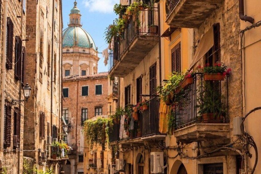A narrow street with buildings, balconies with plants, and a dome in the background. 4