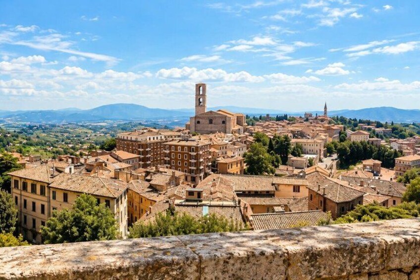 A historic town with a prominent tower, terracotta-roofed buildings, and a mountainous backdrop. 2