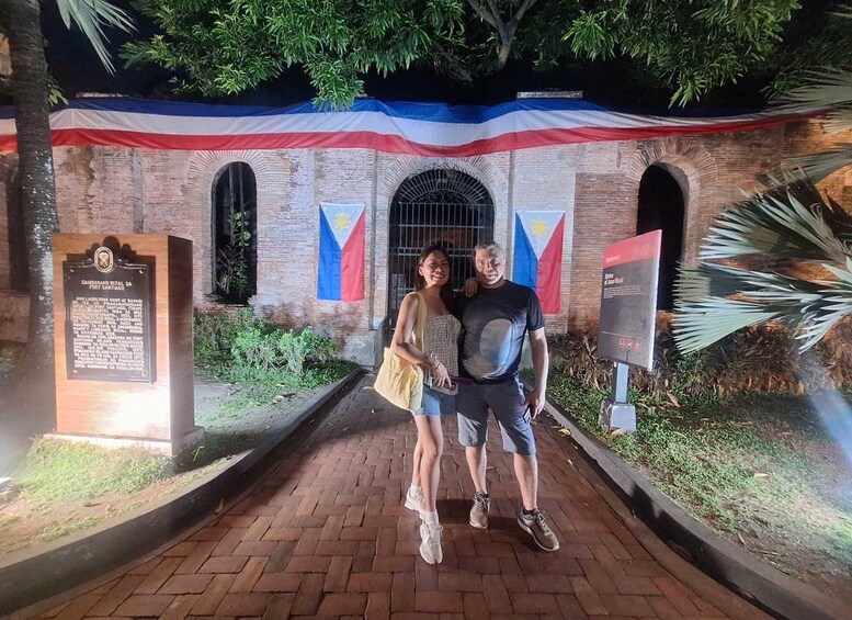 A couple posing in front of a historical building with a Philippine flag and a plaque. 3