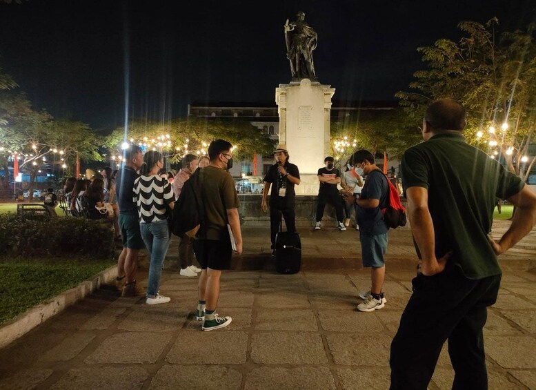 A nighttime outdoor gathering with people standing and conversing, a statue in the background, and trees with string lights. 1
