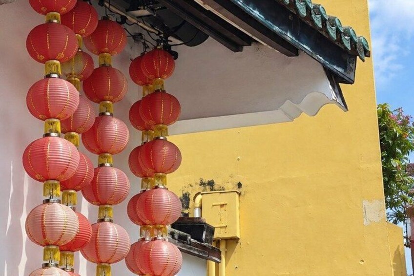 A building with a yellow facade, red lanterns hanging from the roof, and a black electrical box attached to the wall. 3