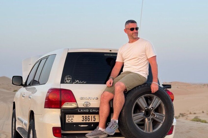 A man sitting on the rear tire of a white SUV in a desert landscape. 1