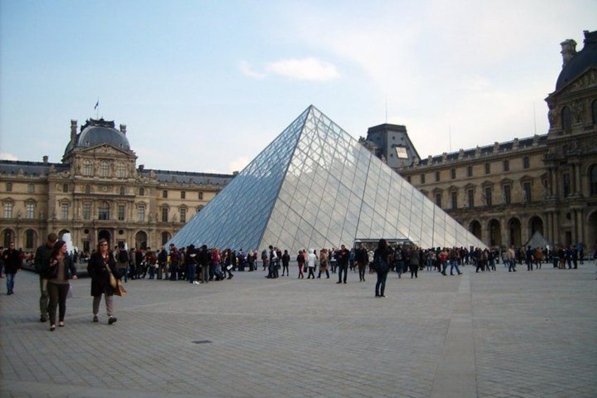 The Louvre Pyramid in the center of a large courtyard with people around it. 1