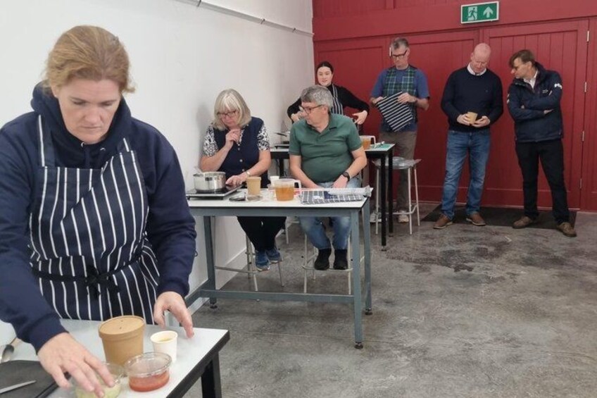 A person in a striped apron working at a table with various kitchen items, surrounded by seated individuals and others standing. 3