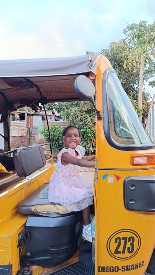 A child sitting in a yellow auto-rickshaw with the number 273, parked in a rural area with greenery and buildings in the background. 2