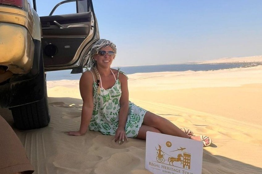 A person sitting on sand dunes with a vehicle and a sign indicating a heritage tour. 1