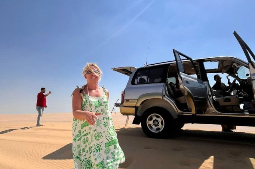 A person in a green patterned dress standing in a desert with a vehicle with its doors open. 5