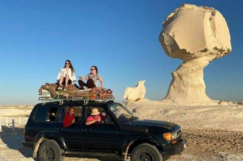 A black SUV with passengers on the roof, a large rock formation, and a smaller rock in the background. 3