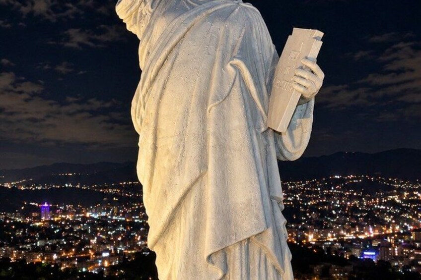 A statue of a person holding a book, set against a cityscape at night. 3