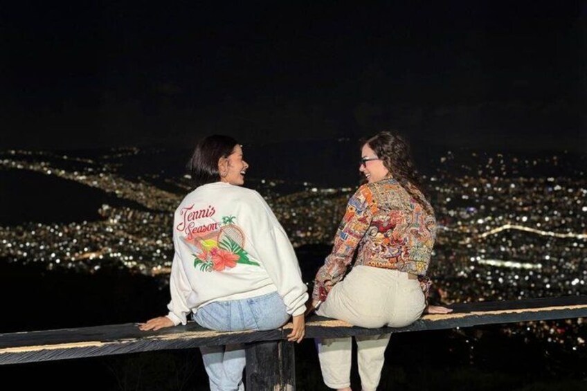 Two people sitting on a railing overlooking a cityscape at night. 1
