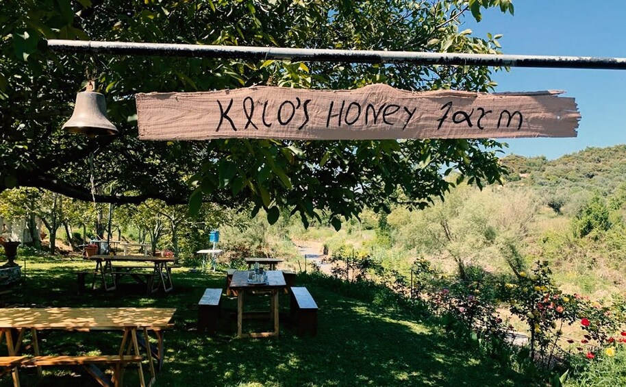 Ein Holzschild mit der Aufschrift 'K&L's Honey Farm', umgeben von Picknicktischen und einer Glocke, inmitten einer üppigen, grünen Landschaft. 2
