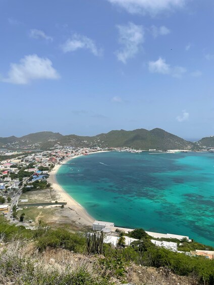 A coastal town with a beach, clear turquoise waters, and green hills in the background. 1