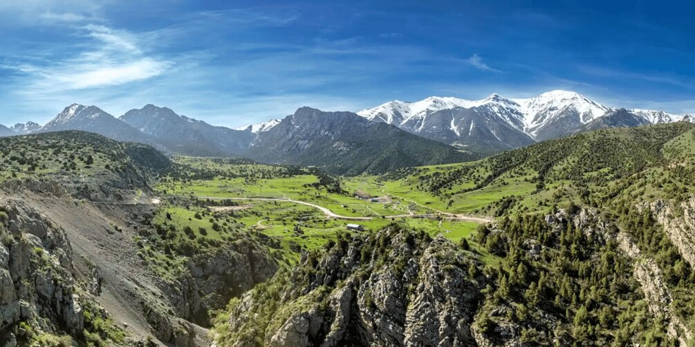 A mountainous landscape with a valley, a winding road, and snow-capped peaks in the background. 2