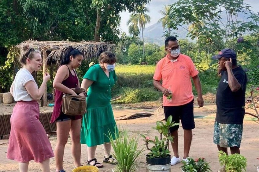 A group of people outdoors, some standing and some sitting, with greenery and a thatched structure in the background. 4
