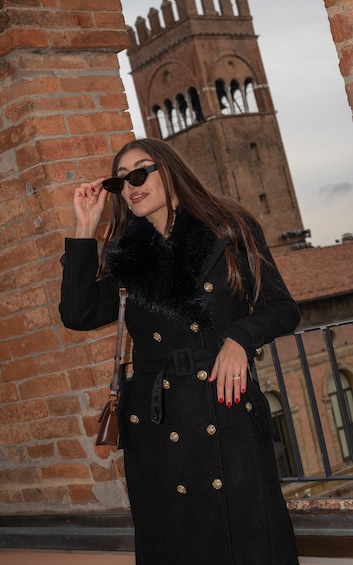 A person wearing a black coat with gold buttons, standing in front of a brick wall with a tower in the background. 5