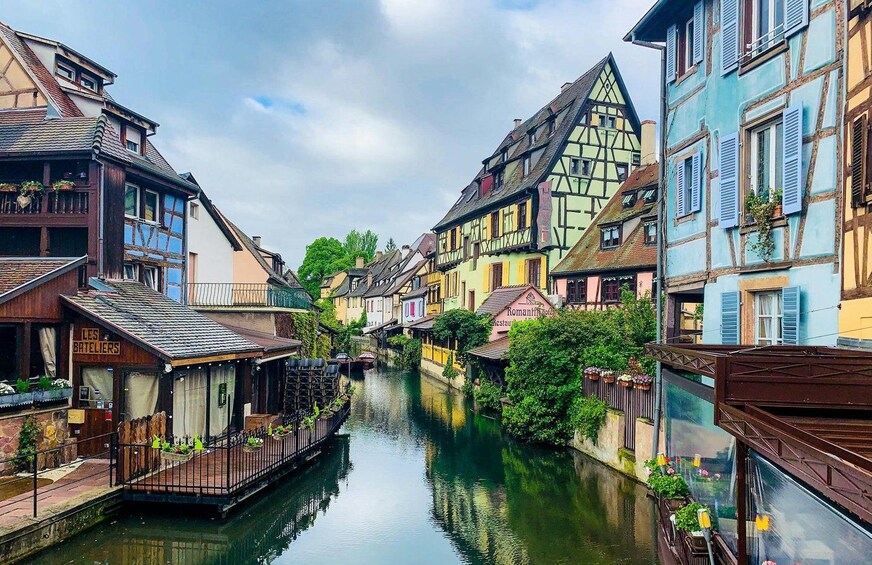 A narrow waterway flanked by colorful half-timbered buildings, a wooden boat docked on the left, and a sign for 'Les Bateliers' restaurant. 2