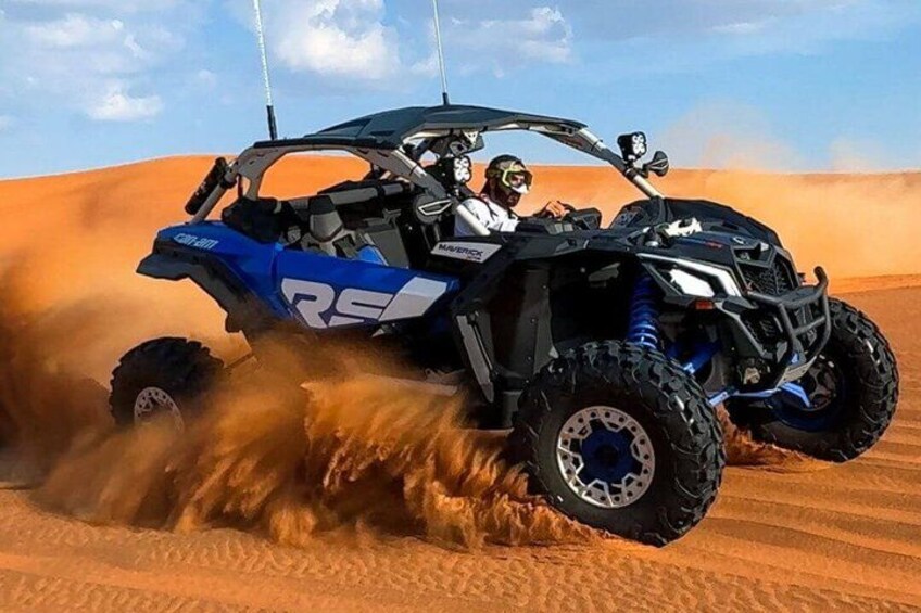 A blue and black ATV with the brand name 'RS' on the side, driving through a desert with sand being kicked up by the tires. 1