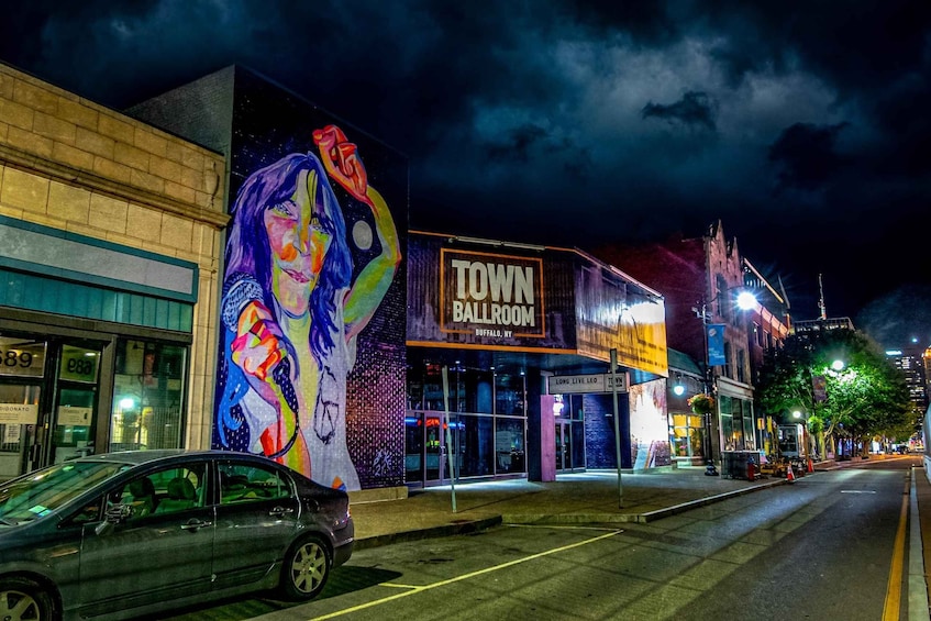 A street at night with a mural on a building, a car parked on the side, and a sign for 'Town Ball Room Buffalo, NY'. 3