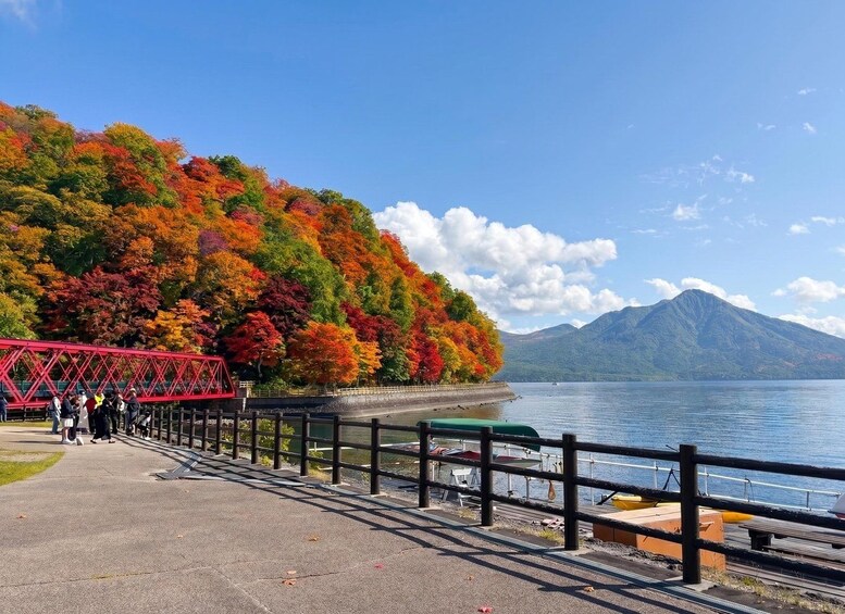 A scenic lakeside walkway with a vibrant autumnal forest, a red bridge, and a mountain in the background. 4
