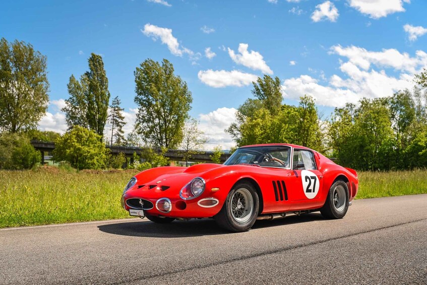 A red vintage sports car with the number 27 on its side, parked on a road with greenery and trees in the background. 4